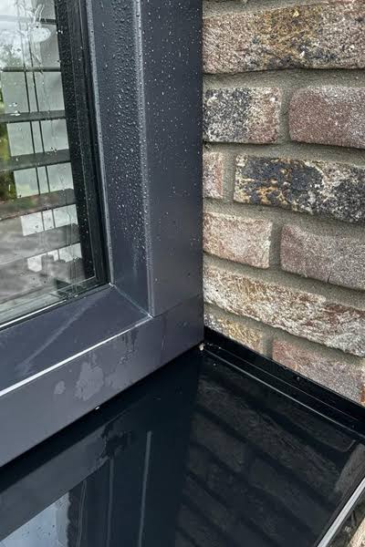 Detail van een donker kozijn met waterdruppels, een natte vensterbank en metselwerk