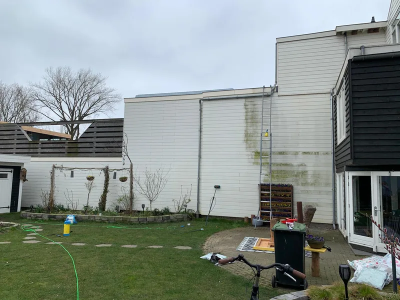 Witte houten gevelbekleding tijdens reiniging, met schone delen links en groene aanslag op het nog te reinigen deel rechts bij de ladder